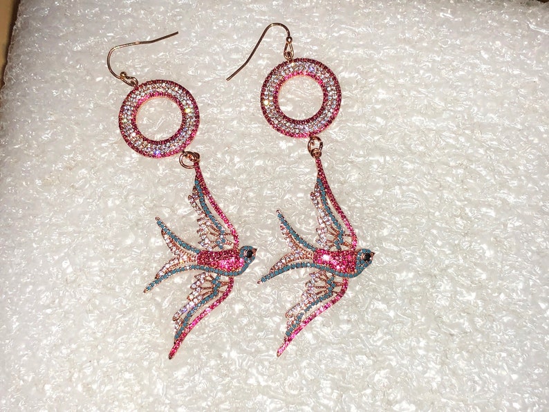 Puede incluir: Un par de pendientes colgantes con un dise&ntilde;o de p&aacute;jaro rosa y azul. Los pendientes tienen un aro redondo engastado con cristales y un colgante de p&aacute;jaro. Los p&aacute;jaros est&aacute;n adornados con cristales rosas y azules.