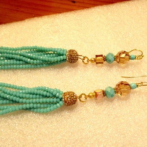 May include: A pair of turquoise beaded tassel earrings. Each earring features a cluster of turquoise beads, a gold-colored rhinestone-studded ball, and a series of gold-colored beads and amber-colored faceted beads. The earrings have gold-colored ear wires.