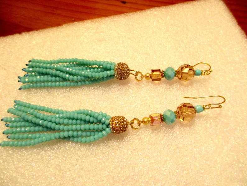May include: A pair of turquoise beaded tassel earrings. Each earring features a cluster of turquoise beads, a gold-colored rhinestone-studded ball, and a series of gold-colored beads and amber-colored faceted beads. The earrings have gold-colored ear wires.