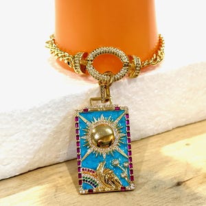 May include: A gold chain necklace with a square pendant featuring a blue enamel background with a gold sun and a rainbow. The pendant is surrounded by pink and white stones.