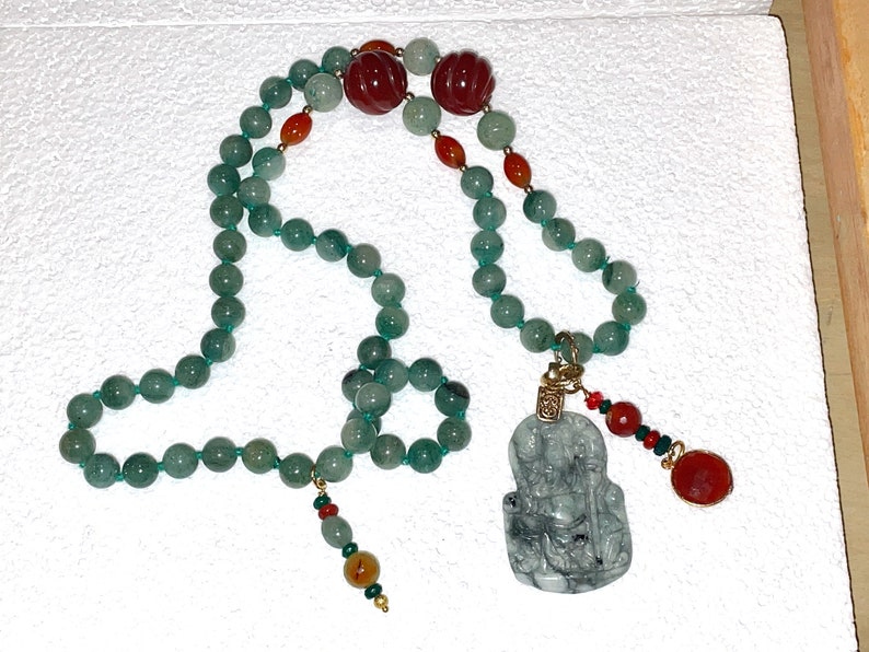 Puede incluir: Un collar de cuentas de jade verde con un colgante de jade con una figura tallada. El collar tambi&eacute;n incluye cuentas rojas y naranjas.