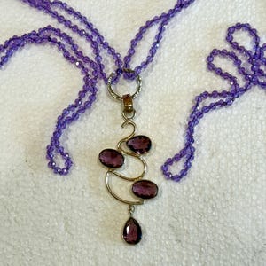 May include: A purple amethyst gemstone necklace with a pendant featuring a silver design with multiple amethyst stones. The necklace is made of multiple strands of small purple beads.