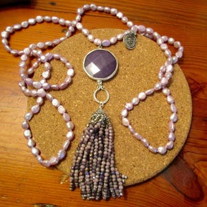 May include: A long lavender pearl necklace with a silver pendant featuring a large purple gemstone and a tassel of smaller purple beads.