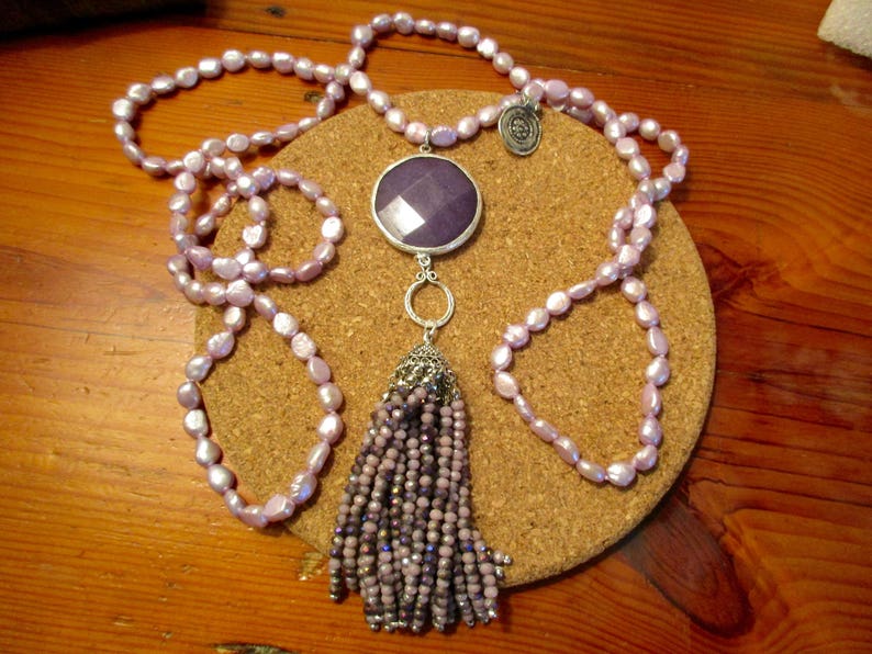 May include: A long lavender pearl necklace with a silver pendant featuring a large purple gemstone and a tassel of smaller purple beads.