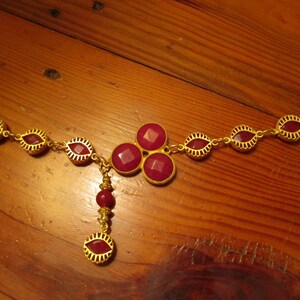 May include: A gold necklace with a pendant featuring a cluster of four red gemstones. The necklace is adorned with smaller red gemstones throughout.