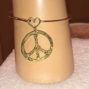 May include: A gold bangle bracelet with a peace sign charm and a heart charm. The peace sign charm has a small heart in the center.