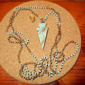 May include: A turquoise and gold arrowhead pendant necklace with a double strand of beads. The necklace is on a cork background.