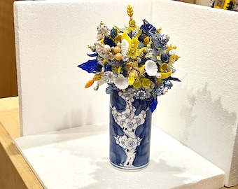 May include: A blue and white ceramic vase with a floral design, holding a bouquet of yellow, white, and blue glass flowers.