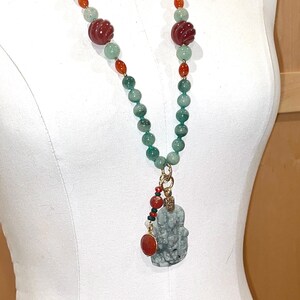 Puede incluir: Un collar largo de cuentas verdes y naranjas con un colgante de jade tallado. El colgante est&aacute; unido al collar con una cadena de oro y tiene una cuenta roja en la parte superior.