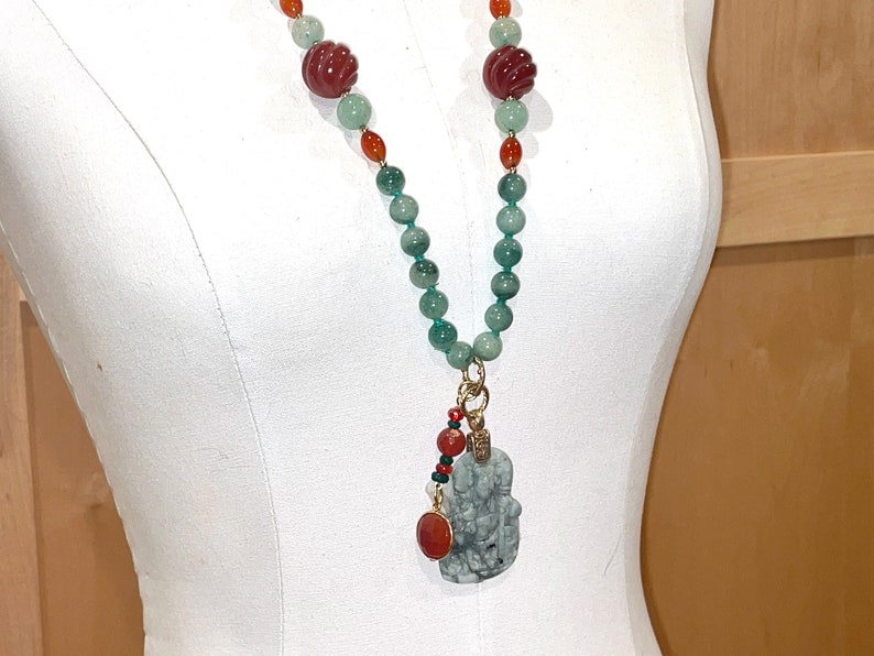 Puede incluir: Un collar largo de cuentas verdes y naranjas con un colgante de jade tallado. El colgante est&aacute; unido al collar con una cadena de oro y tiene una cuenta roja en la parte superior.