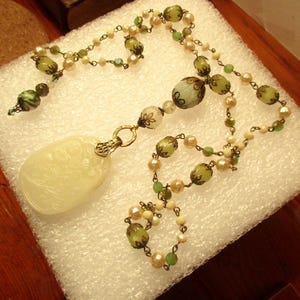 Puede incluir: Un collar largo y delicado con un colgante de jade blanco tallado. El collar presenta cuentas de vidrio verde y blanco y detalles en tono dorado.