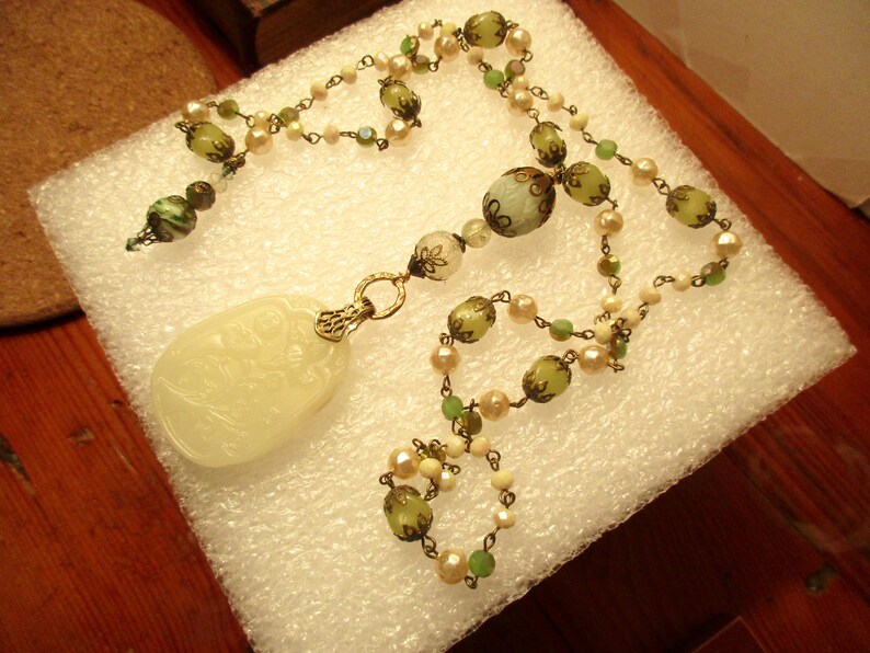 Puede incluir: Un collar largo y delicado con un colgante de jade blanco tallado. El collar presenta cuentas de vidrio verde y blanco y detalles en tono dorado.