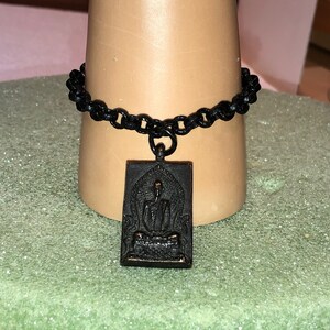 May include: A black chain bracelet with a black metal pendant featuring a raised image of a seated Buddha.