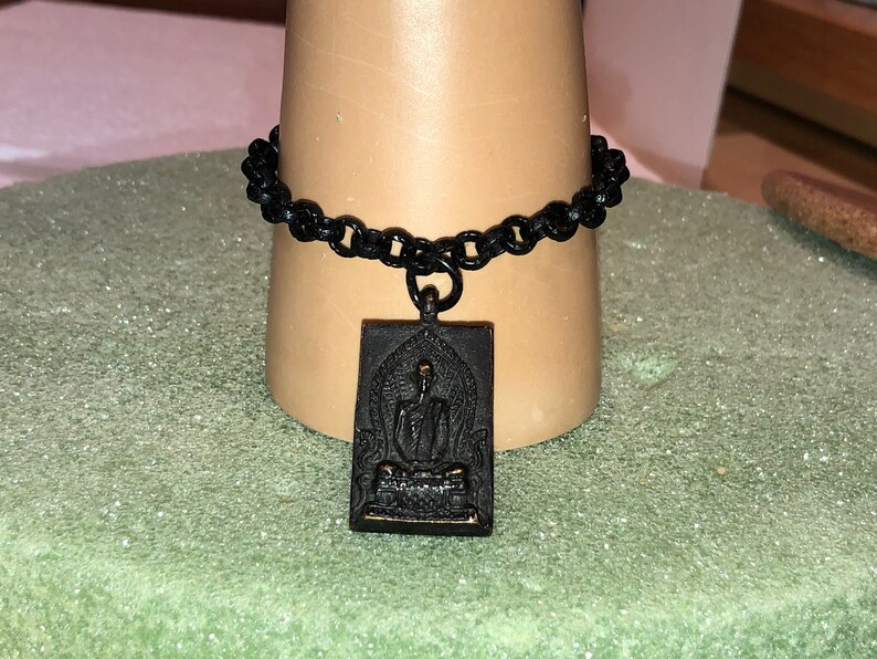 May include: A black chain bracelet with a black metal pendant featuring a raised image of a seated Buddha.