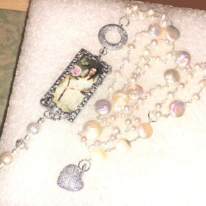 Pu&ograve; includere: Una collana d'argento con un ciondolo rettangolare che presenta un'illustrazione vintage di una donna che tiene un fiore. La collana &egrave; realizzata con pi&ugrave; fili di perle bianche e un ciondolo a forma di cuore.