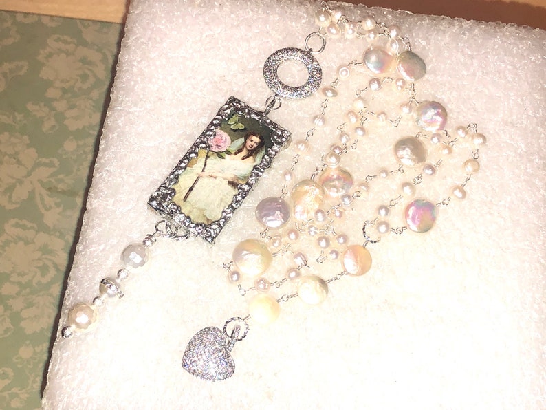 Pu&ograve; includere: Una collana d'argento con un ciondolo rettangolare che presenta un'illustrazione vintage di una donna che tiene un fiore. La collana &egrave; realizzata con pi&ugrave; fili di perle bianche e un ciondolo a forma di cuore.