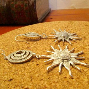 May include: A pair of silver earrings with a sunburst design. The earrings are made of metal and have a sparkly finish. The earrings are on a cork surface.