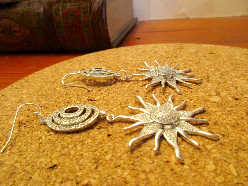 May include: A pair of silver earrings with a sunburst design. The earrings are made of metal and have a sparkly finish. The earrings are on a cork surface.
