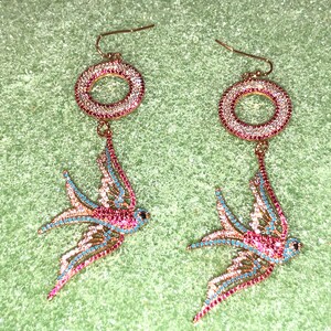 Puede incluir: Un par de pendientes de tono dorado con un dise&ntilde;o de p&aacute;jaro colgante. Los p&aacute;jaros est&aacute;n hechos de pedrer&iacute;a transparente, rosa y azul. Los pendientes tambi&eacute;n presentan un dise&ntilde;o circular con pedrer&iacute;a.