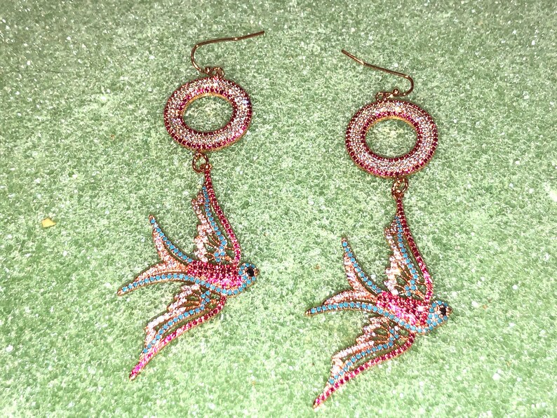 Puede incluir: Un par de pendientes de tono dorado con un dise&ntilde;o de p&aacute;jaro colgante. Los p&aacute;jaros est&aacute;n hechos de pedrer&iacute;a transparente, rosa y azul. Los pendientes tambi&eacute;n presentan un dise&ntilde;o circular con pedrer&iacute;a.