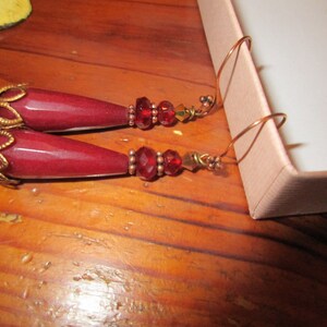 May include: A pair of dangle earrings with burgundy teardrop-shaped stones, gold accents, and red crystal beads.