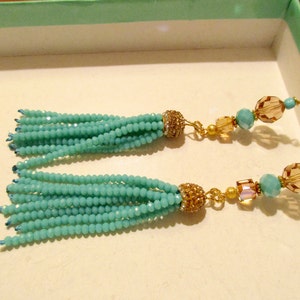 May include: A pair of turquoise beaded tassel earrings. The earrings feature multiple strands of small, round turquoise beads, gold-colored accents, and decorative gold-colored bead caps. The earrings have gold-colored ear wires.