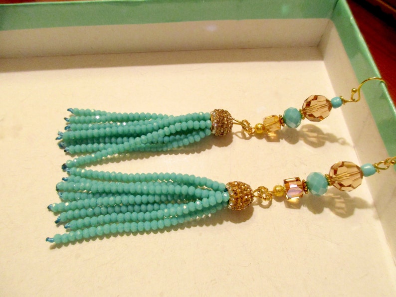 May include: A pair of turquoise beaded tassel earrings. The earrings feature multiple strands of small, round turquoise beads, gold-colored accents, and decorative gold-colored bead caps. The earrings have gold-colored ear wires.