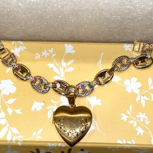 May include: A gold chain bracelet with a heart-shaped locket. The chain is decorated with colorful gemstones. The locket has a floral design.