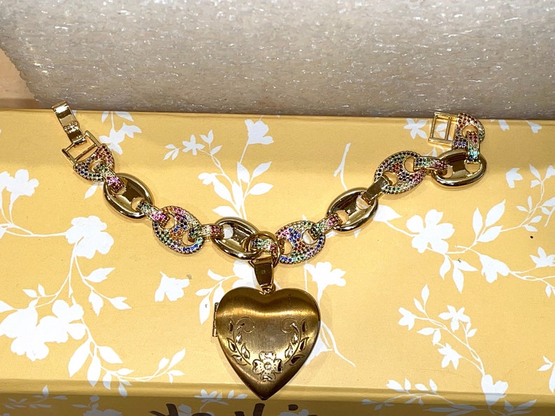 May include: A gold chain bracelet with a heart-shaped locket. The chain is decorated with colorful gemstones. The locket has a floral design.