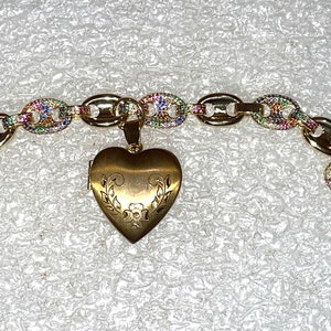 May include: A gold chain bracelet with a heart-shaped locket. The chain is decorated with colorful gemstones. The locket has a floral design.