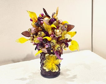May include: A purple glass vase with a textured surface is filled with a colorful arrangement of yellow and purple glass flowers and beads.