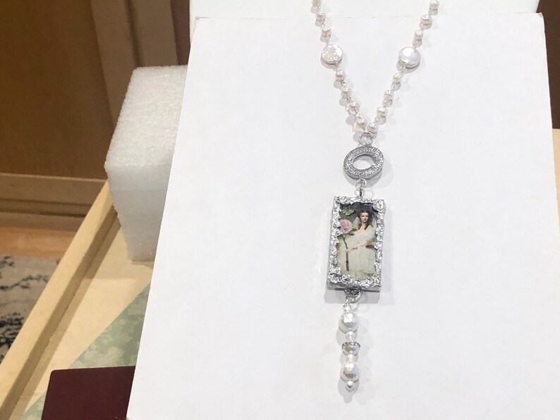 Pu&ograve; includere: Una collana d'argento con un ciondolo rettangolare che presenta un ritratto vintage di una donna. Il ciondolo &egrave; circondato da piccole pietre trasparenti. La collana &egrave; realizzata con perle bianche e argento.