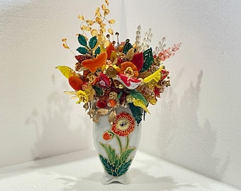 May include: A white ceramic vase with a floral design and a bouquet of colourful glass flowers. The flowers are made of glass and have a variety of shapes and sizes. The vase is decorated with a green and gold floral pattern.