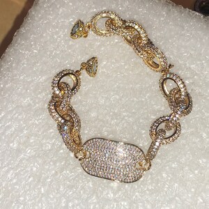 May include: A gold bracelet with a large, rectangular charm covered in sparkling stones. The bracelet is made of linked chains, each covered in small, clear stones.