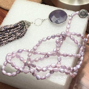 May include: A long lavender pearl necklace with a large amethyst gemstone pendant and a beaded tassel.