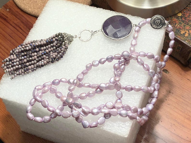 May include: A long lavender pearl necklace with a large amethyst gemstone pendant and a beaded tassel.