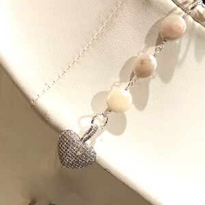 Pu&ograve; includere: Una collana a catena d'argento con un ciondolo a forma di cuore incrostato di piccole pietre trasparenti. La collana presenta una serie di perle rotonde di colore avorio.