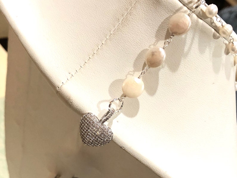 Pu&ograve; includere: Una collana a catena d'argento con un ciondolo a forma di cuore incrostato di piccole pietre trasparenti. La collana presenta una serie di perle rotonde di colore avorio.