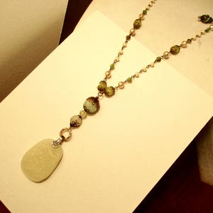 Puede incluir: Un collar largo con cuentas verdes, blancas y marrones. El collar tiene un colgante ovalado grande de color blanco.