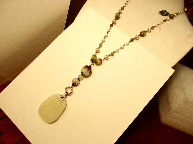 Puede incluir: Un collar largo con cuentas verdes, blancas y marrones. El collar tiene un colgante ovalado grande de color blanco.