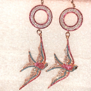 Puede incluir: Un par de pendientes de tono dorado con un dise&ntilde;o circular y un colgante de p&aacute;jaro golondrina. Los pendientes est&aacute;n adornados con cristales rosas y azules.