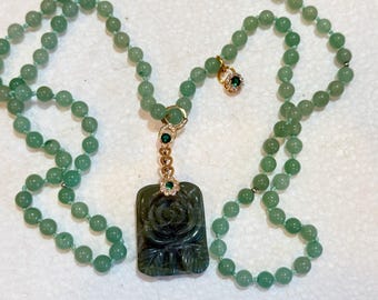 May include: A green beaded necklace with a carved dark green pendant featuring a rose design. The pendant is accented with gold and emerald-colored gemstones. The necklace has a gold clasp and a small charm with a green gemstone.