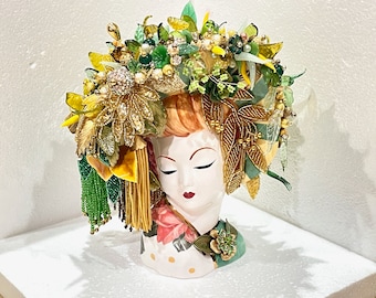May include: A ceramic bust of a woman with closed eyes, wearing a crown of green, gold, and white beads and leaves. The crown is adorned with rhinestones and pearls.