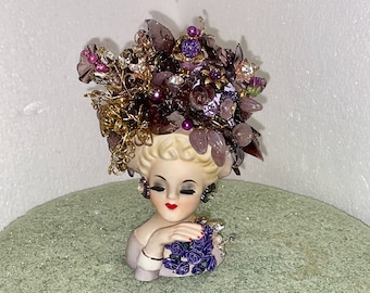 May include: A porcelain figurine of a woman's head with a large, elaborate hairpiece made of purple, brown, and gold embellishments. The hairpiece is adorned with beads, rhinestones, and other decorative elements. The woman is wearing a purple dress and has a bouquet of purple flowers in her hand.