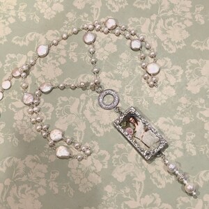 Pu&ograve; includere: Una collana d'argento con un grande ciondolo rettangolare che presenta un'immagine vintage di una donna con un abito bianco. Il ciondolo &egrave; circondato da strass e pende da una catena d'argento con un ciondolo a forma di cuore. La collana &egrave; realizzata con perle bianche e perline d'argento.