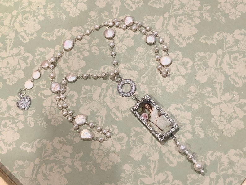 Pu&ograve; includere: Una collana d'argento con un grande ciondolo rettangolare che presenta un'immagine vintage di una donna con un abito bianco. Il ciondolo &egrave; circondato da strass e pende da una catena d'argento con un ciondolo a forma di cuore. La collana &egrave; realizzata con perle bianche e perline d'argento.