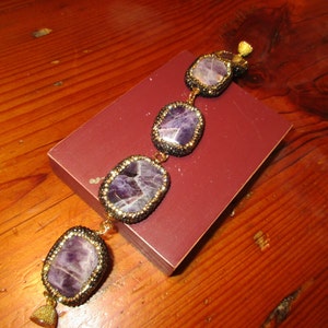 May include: A purple amethyst gemstone bracelet with gold accents and black beads. The bracelet has four large, rectangular amethyst stones.