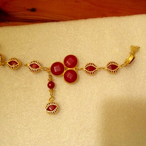 May include: A gold necklace with a red gemstone pendant and red gemstone accents. The necklace has a dangling red gemstone charm.