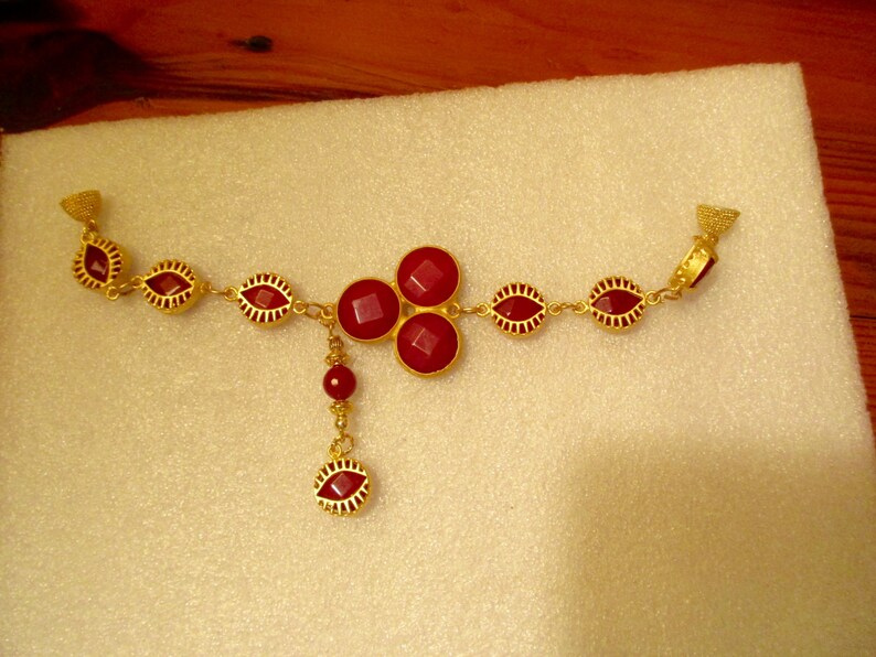 May include: A gold necklace with a red gemstone pendant and red gemstone accents. The necklace has a dangling red gemstone charm.