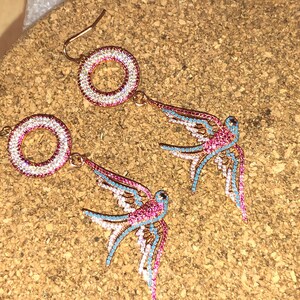 Puede incluir: Un par de pendientes de tono dorado con un dise&ntilde;o circular y un p&aacute;jaro golondrina colgante. Los pendientes est&aacute;n adornados con pedrer&iacute;a rosa, azul y blanca.
